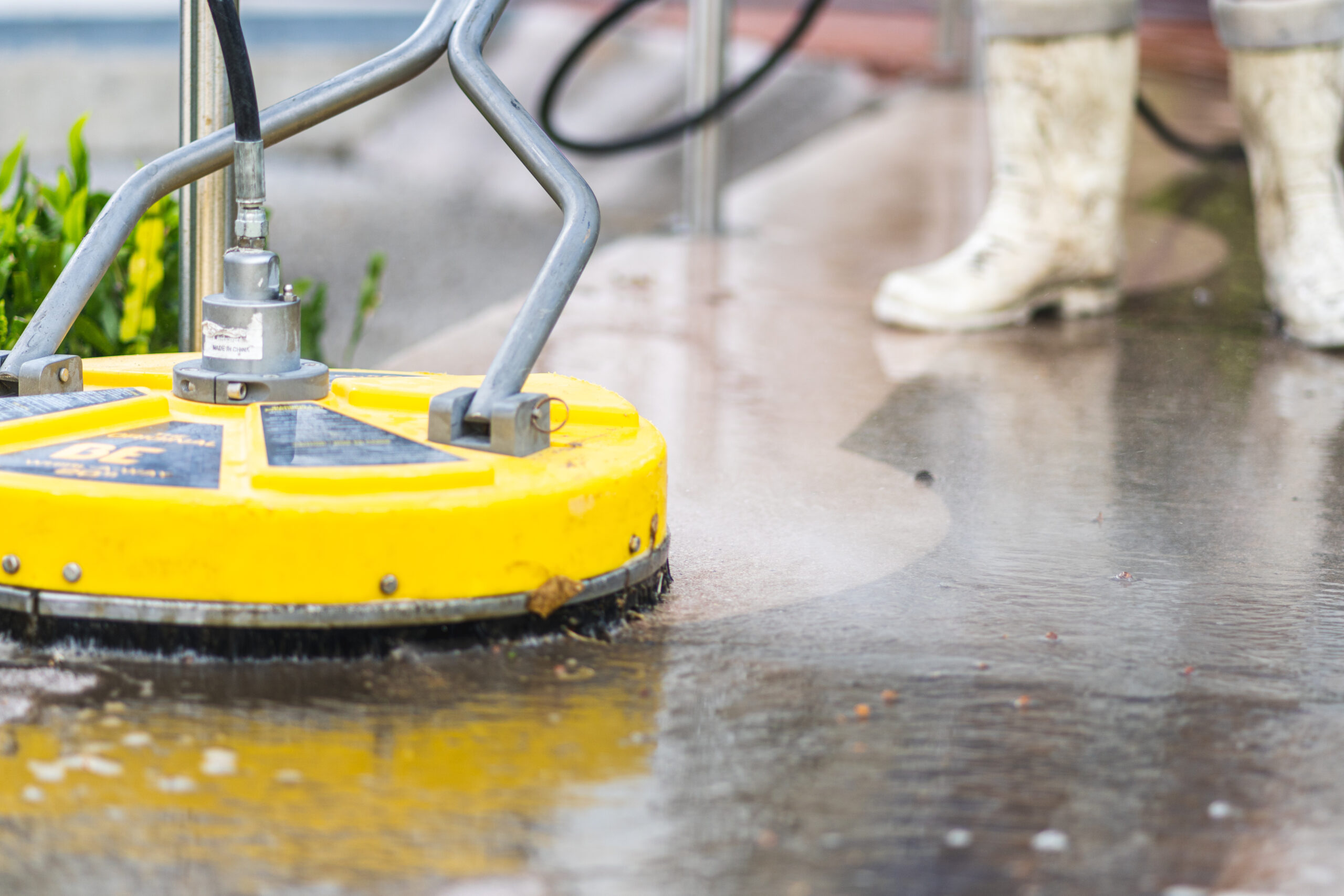 Professional pressure cleaning service in action, with a focus on a high-pressure washer cleaning a concrete driveway, showcasing powerful water jets and a worker wearing dirty white boots