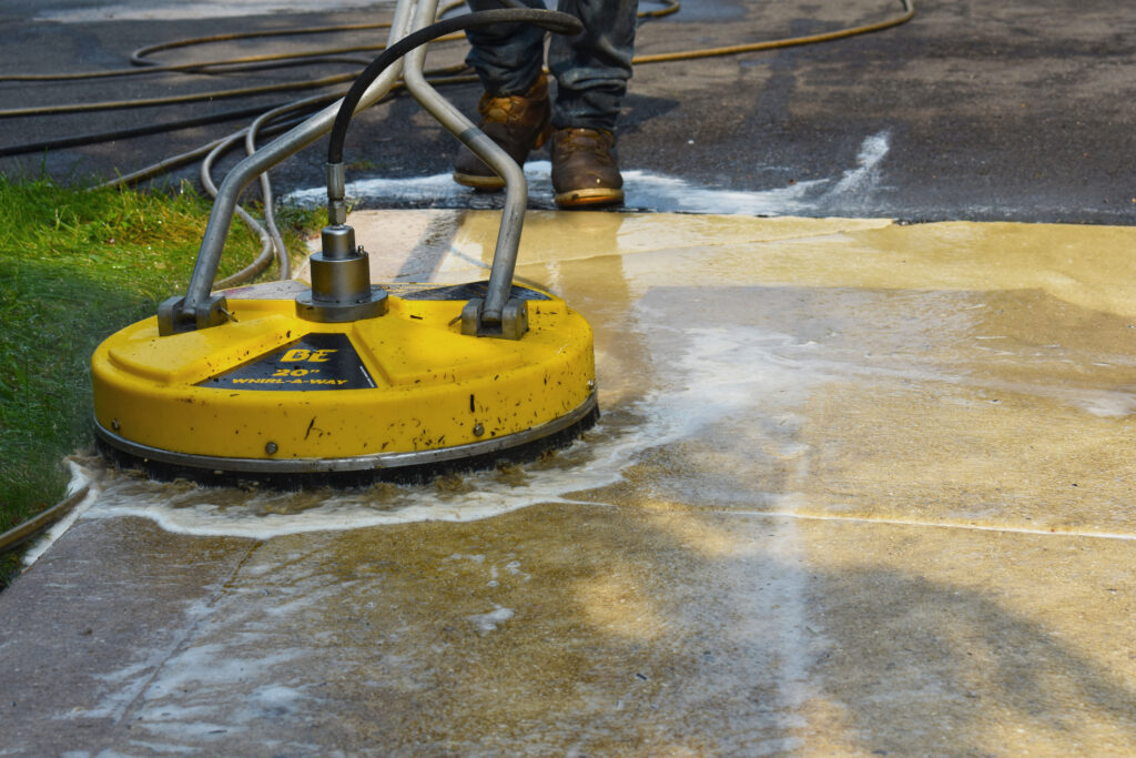 Industrial pressure cleaning in action, featuring a commercial-grade yellow pressure washer cleaning a concrete surface, with water forcefully ejecting and visibly removing dirt.